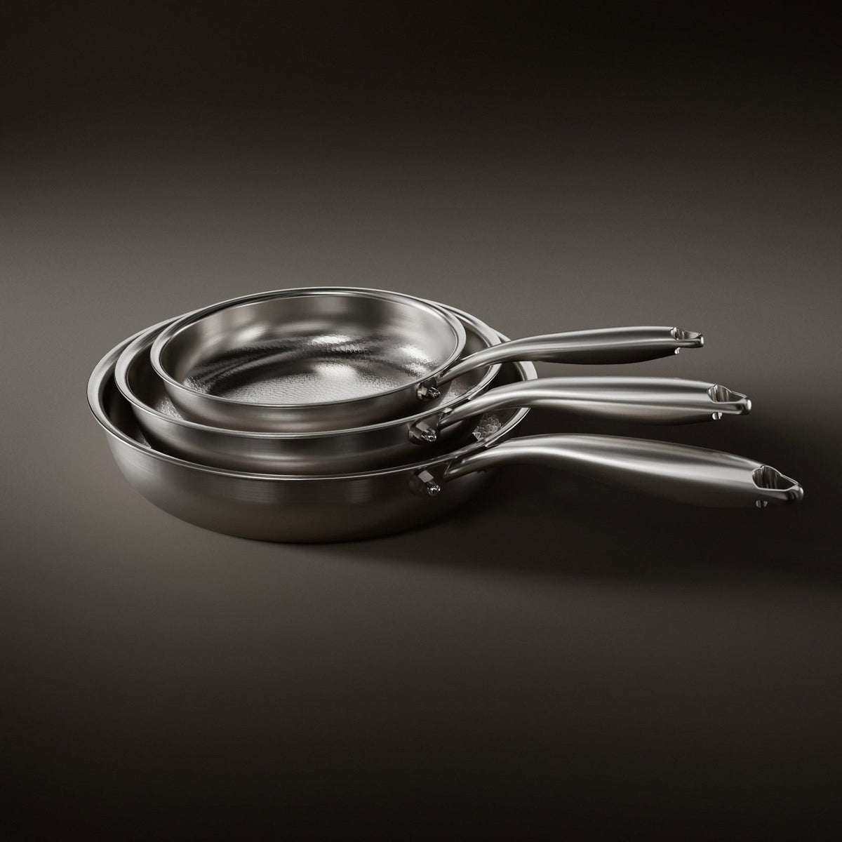 Three hammered metal frying pans nested together by size on a dark background, handles fanned out 1