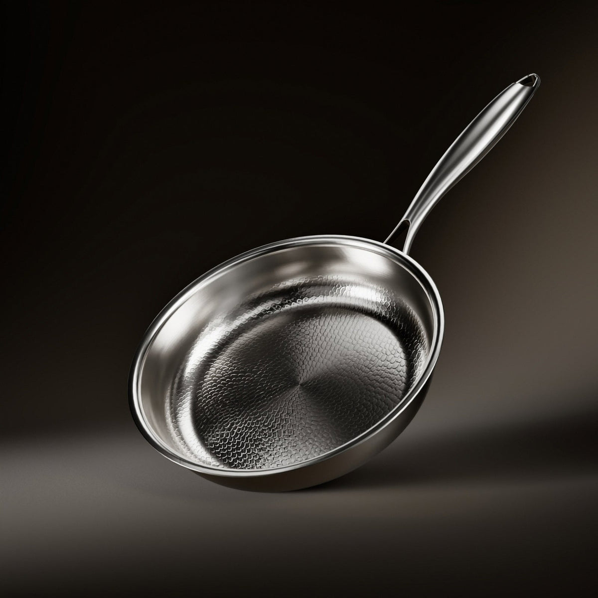 Frying pan tilted against a dark background, showing the hammered cooking surface, polished rim and long stainless steel handle. 1