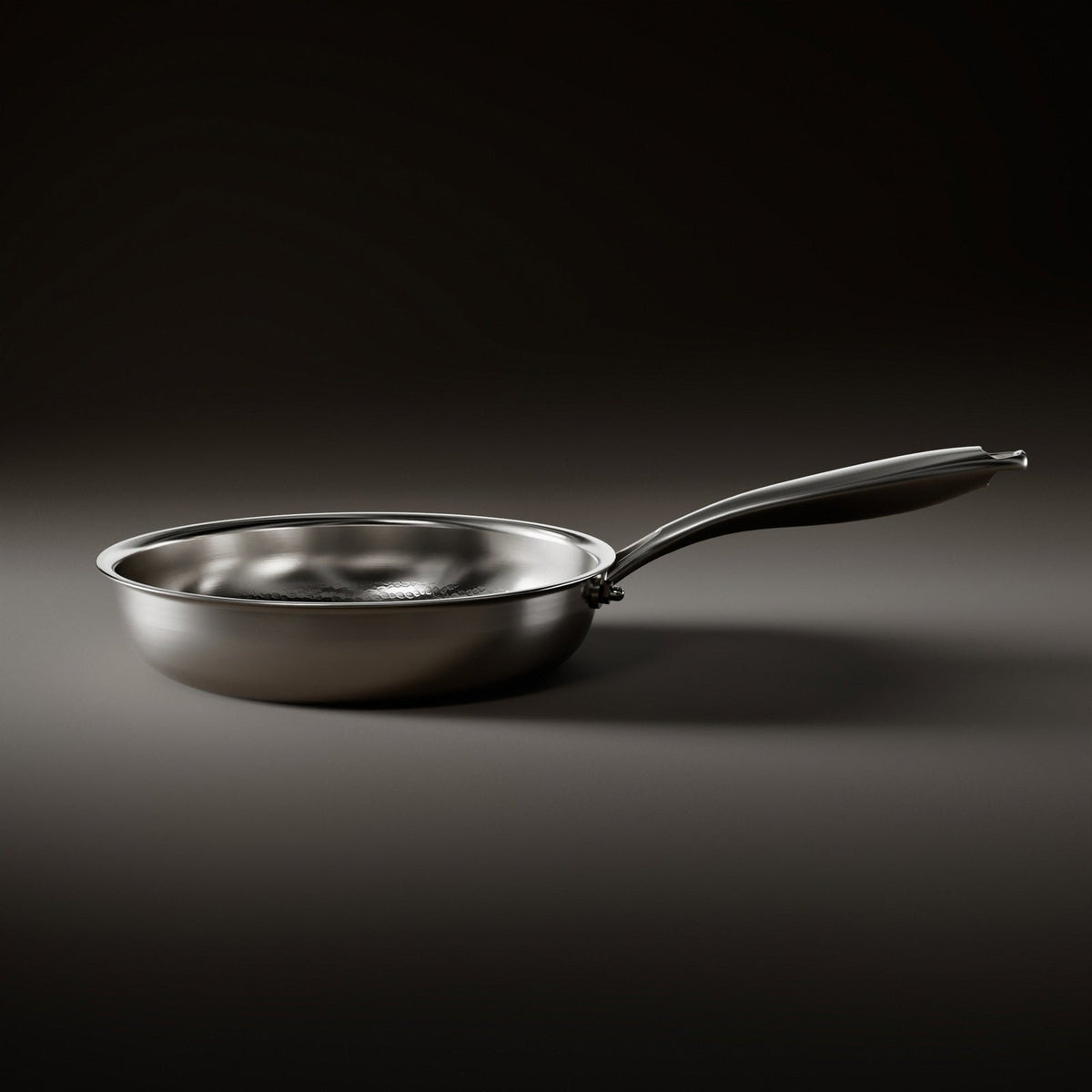 Side view of the frying pan standing flat, showing its shallow curved wall, riveted handle and brushed metal exterior. 2