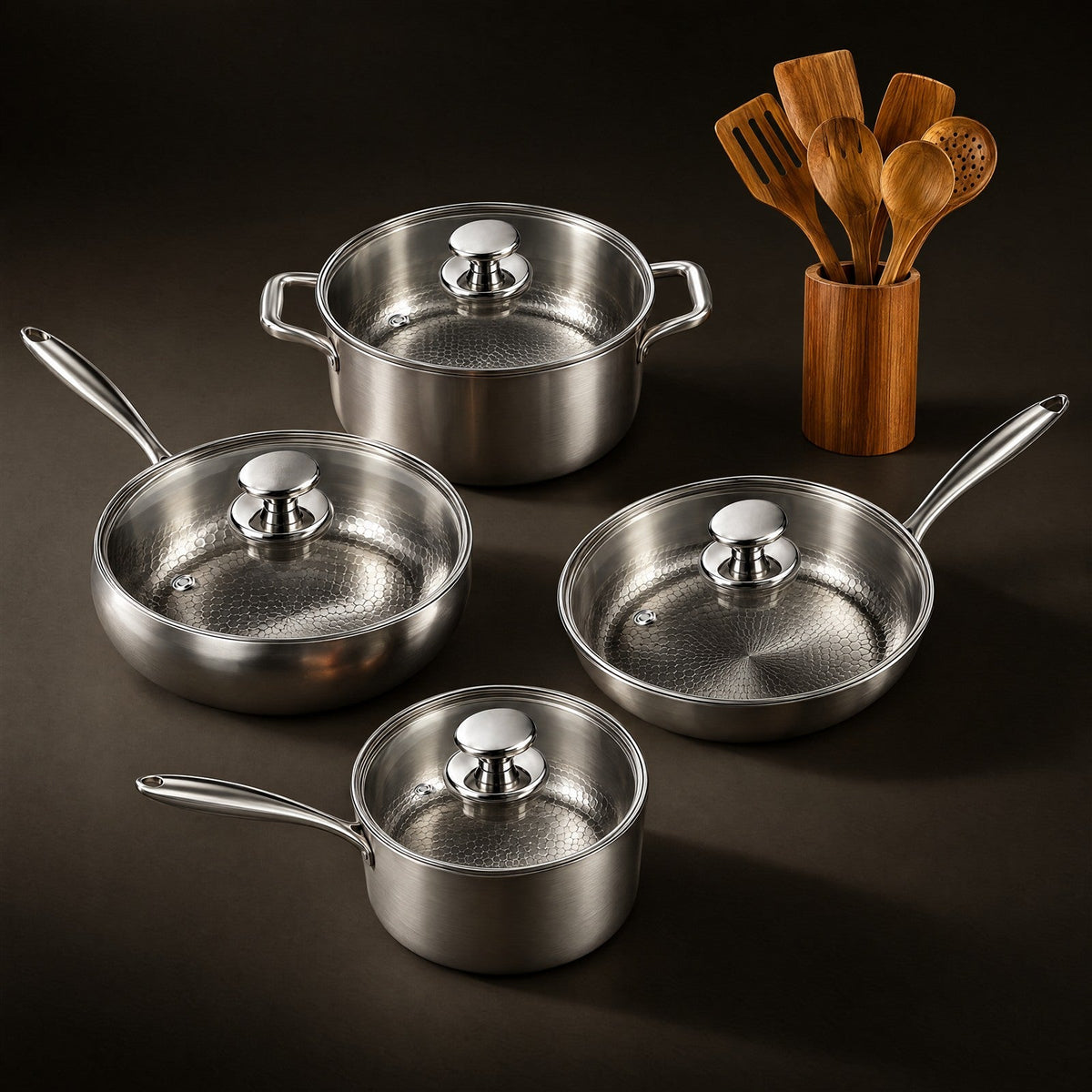 Four lidded hammered titanium pots and pans beside a wooden holder of teak utensils, on a dark surface 1