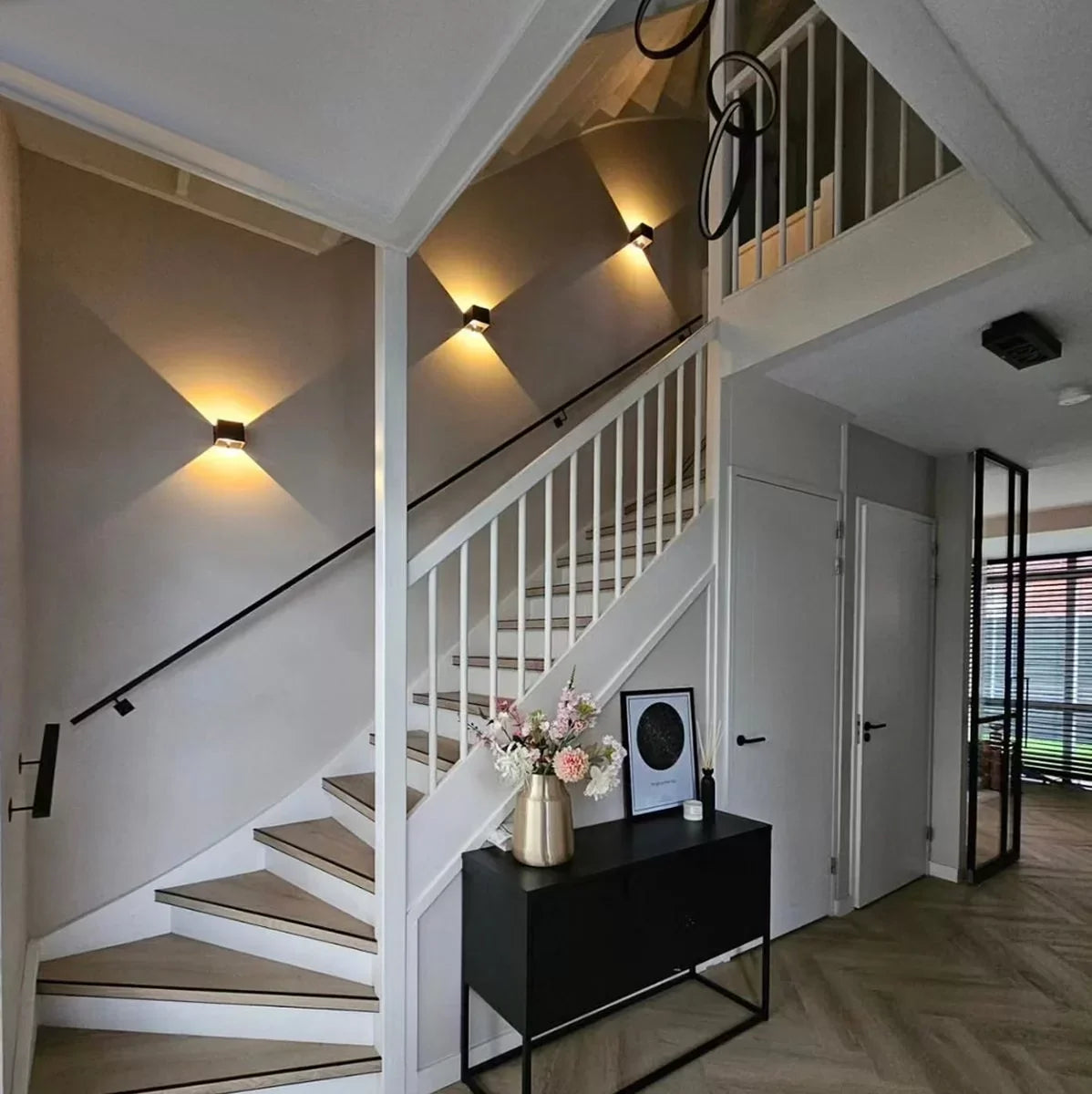 Three black cube wall lamps stepping up a staircase wall, casting warm light above a hallway console table with flowers. 7
