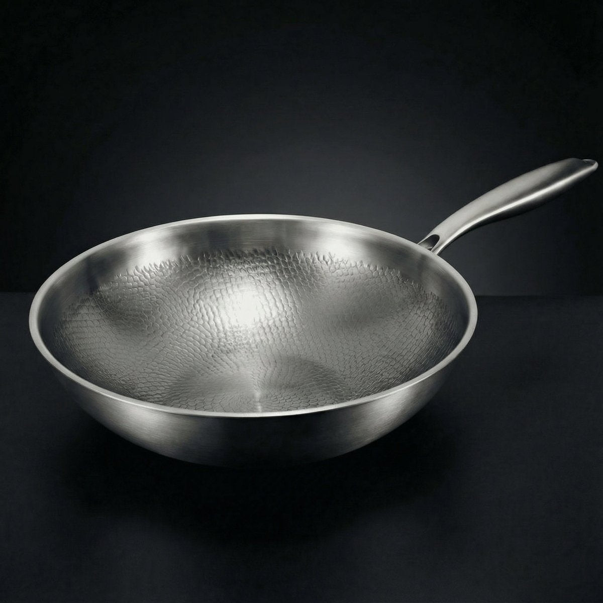 Empty wok seen from a low side angle on a dark surface, showing its hammered interior and long steel handle. 1