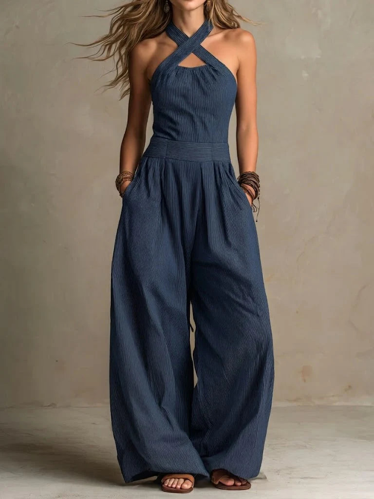Model wearing the crossover-strap jumpsuit in navy blue fine stripes, with a fitted waistband, wide legs, pockets and flat sandals. 3