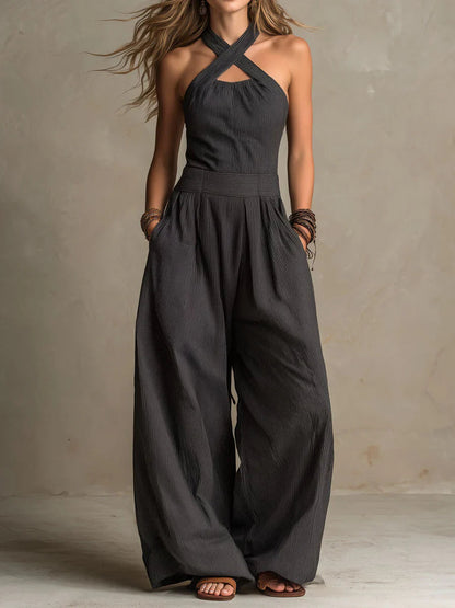Model wearing the wide-leg jumpsuit in dark charcoal pinstripe, with crossed halter straps, hands in pockets, beaded bracelets and tan sandals. 1