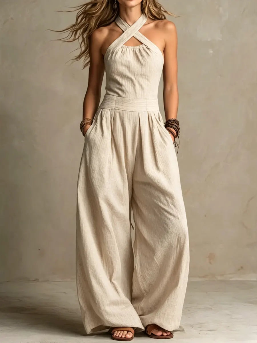 Model wearing the halter jumpsuit in a light cream, textured fabric, with wide flowing legs, hands in pockets and brown sandals. 2