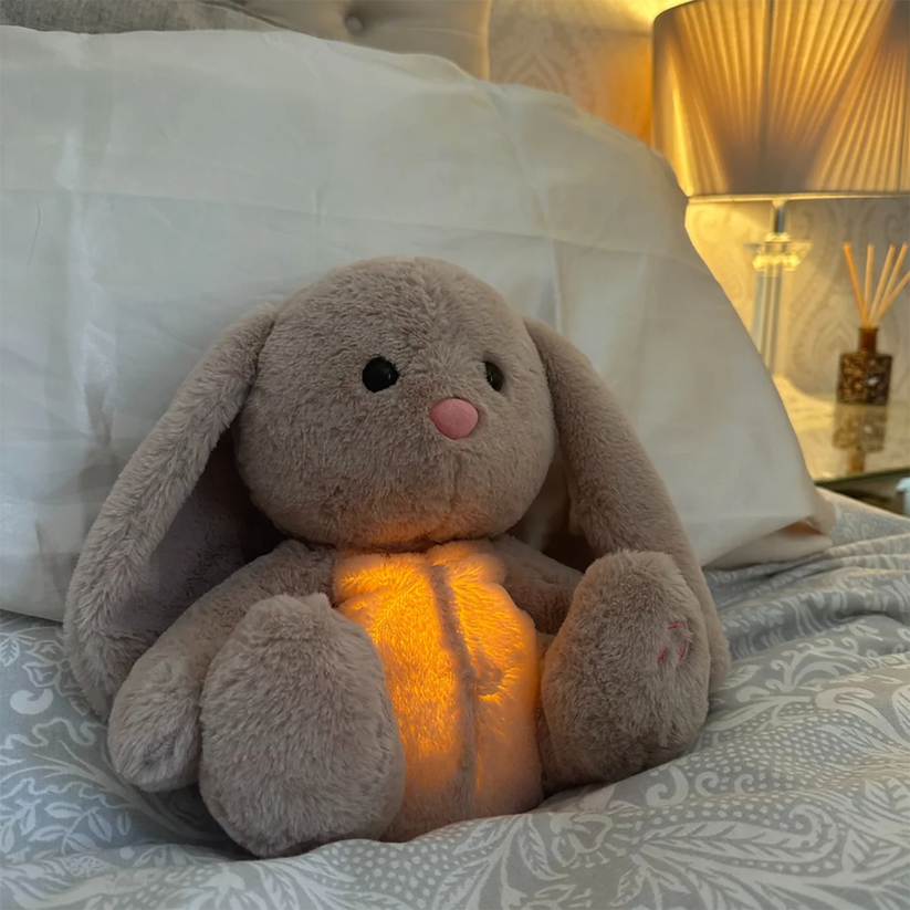 Grey bunny plush with glowing amber tummy propped against a white pillow on a patterned duvet, beside a lit lamp. 15