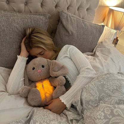 Blonde woman asleep in bed, cuddling a grey bunny plush with a glowing tummy under a patterned duvet. 16