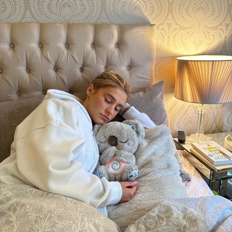 Young woman in a white hoodie asleep in bed, hugging a grey koala plush with a softly lit swirl on its tummy. 14