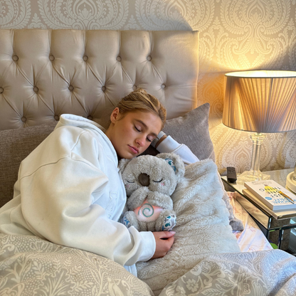 Young woman in a white hoodie asleep in bed, hugging a grey koala plush with a softly lit swirl on its tummy. 14