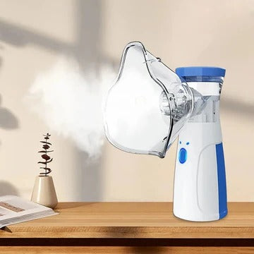 White and blue handheld nebuliser with clear face mask attached, releasing mist, standing on a wooden shelf beside a book and small plant. 1
