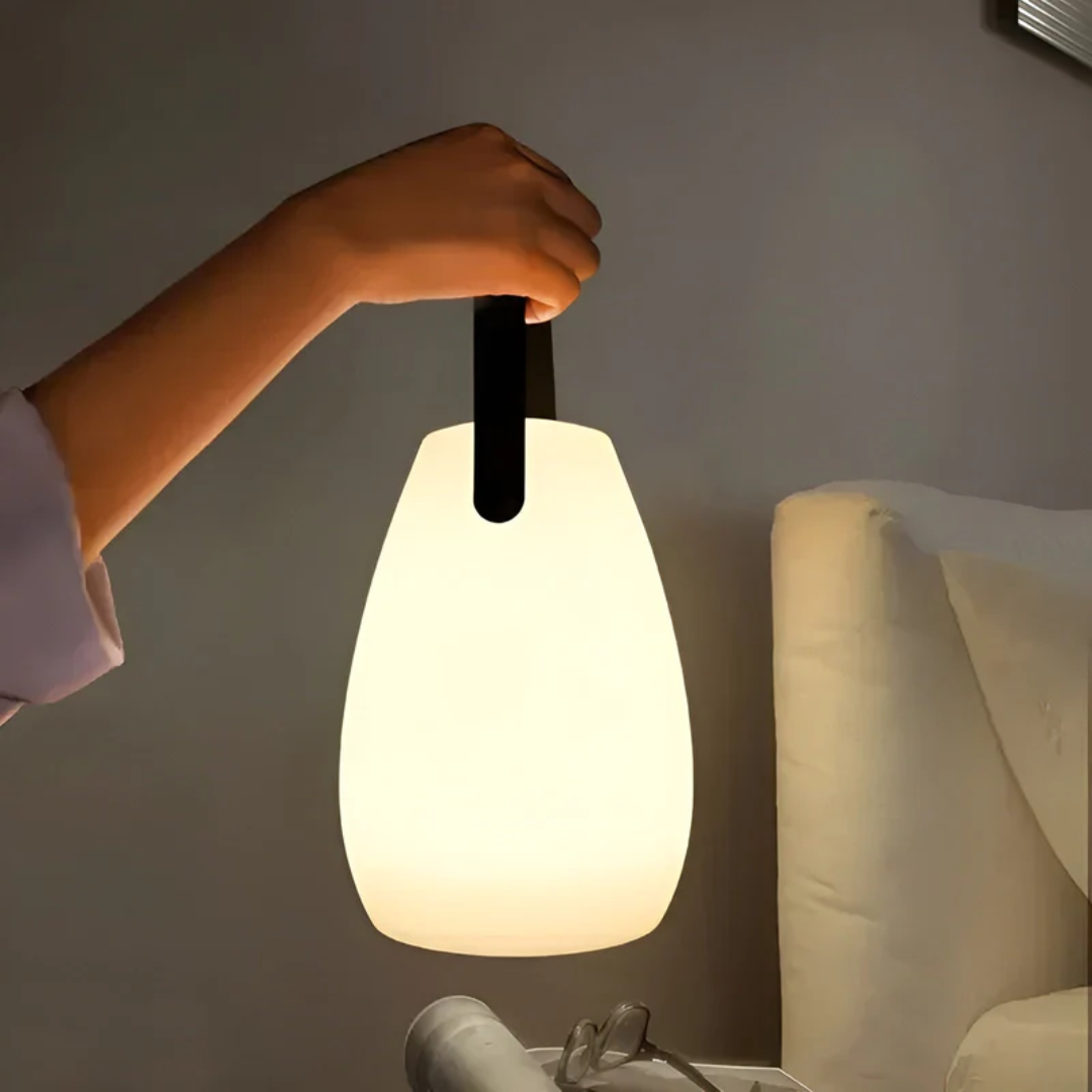 Hand carrying the glowing teardrop lamp by its black strap beside a bed at night 1