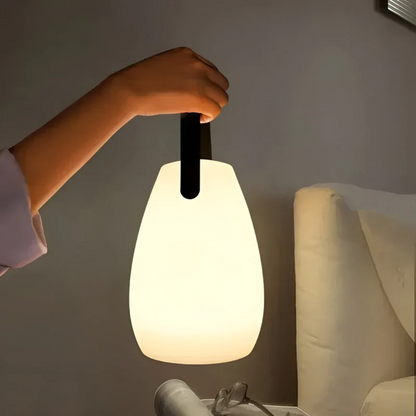 Hand carrying the glowing teardrop lamp by its black strap beside a bed at night 1