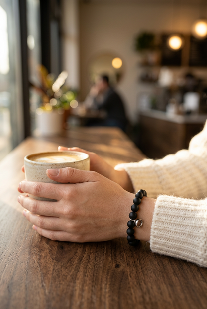 Black beaded bracelet with a small silver charm worn on a wrist holding a latte in a café 1