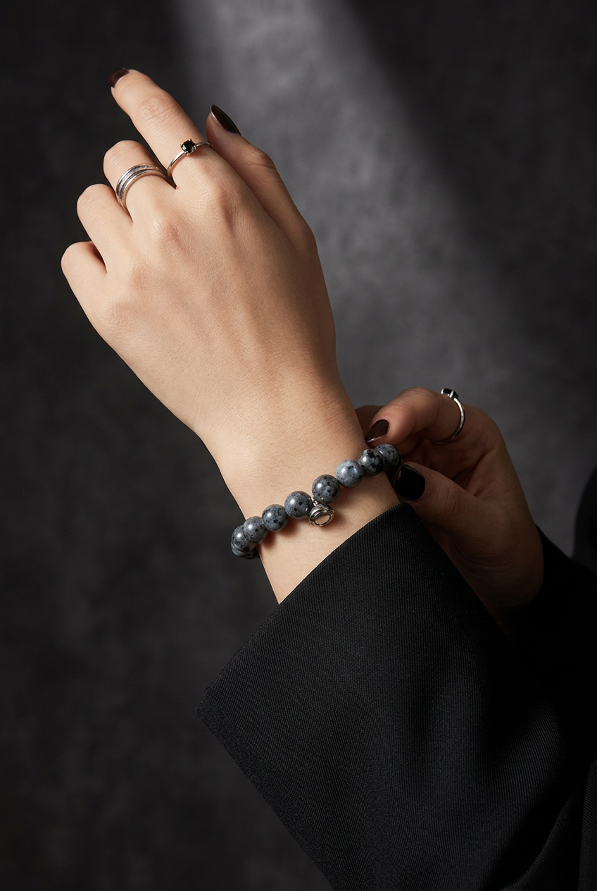 Woman's wrist wearing the grey speckled bead bracelet, with silver rings and a black sleeve against a dark backdrop 4
