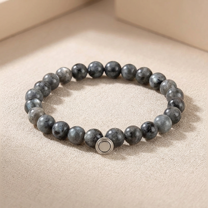 Grey speckled stone bead bracelet with a round silver magnetic clasp, resting on a textured beige surface 3