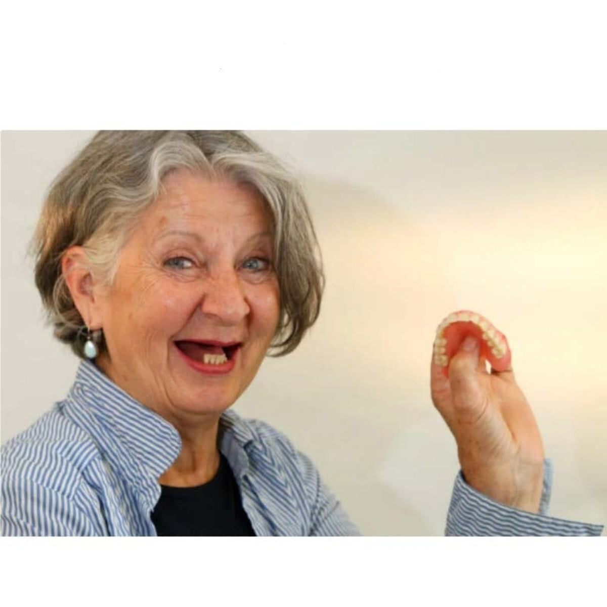 Smiling grey-haired woman in a striped shirt, missing most of her upper teeth, holds up a flexible pink denture. 3