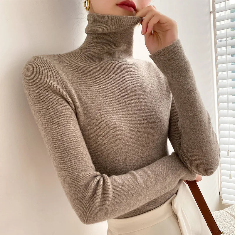 Model wearing the fine knit roll neck jumper in taupe, folding the collar down with one hand, beside a window blind 6