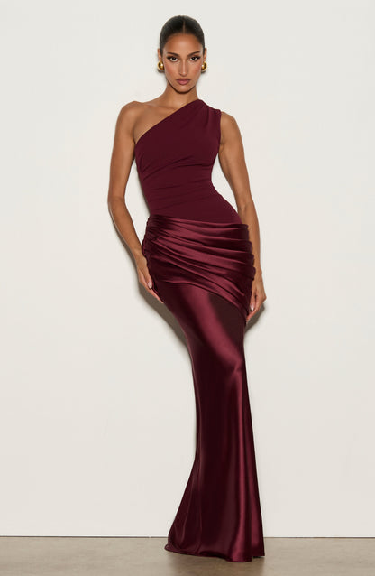 Model posing against a plain wall in the burgundy dress with hand on the hip, showing the fitted shape and satin skirt. 16