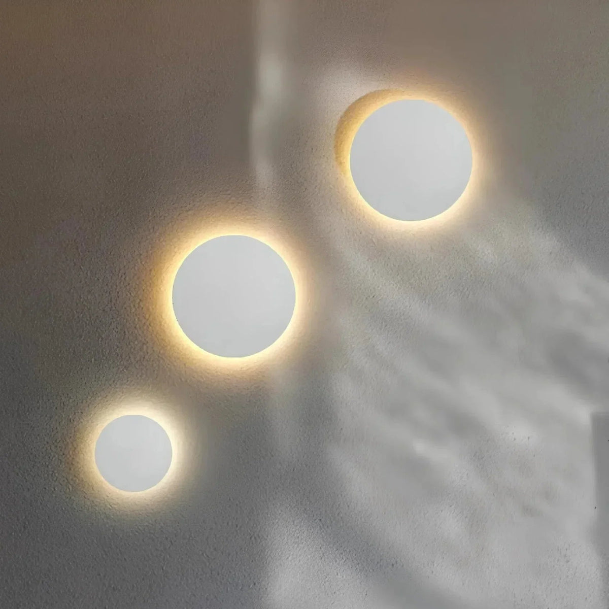 Three white round disc lights in graduated sizes giving off a warm halo on a grey textured wall. 4