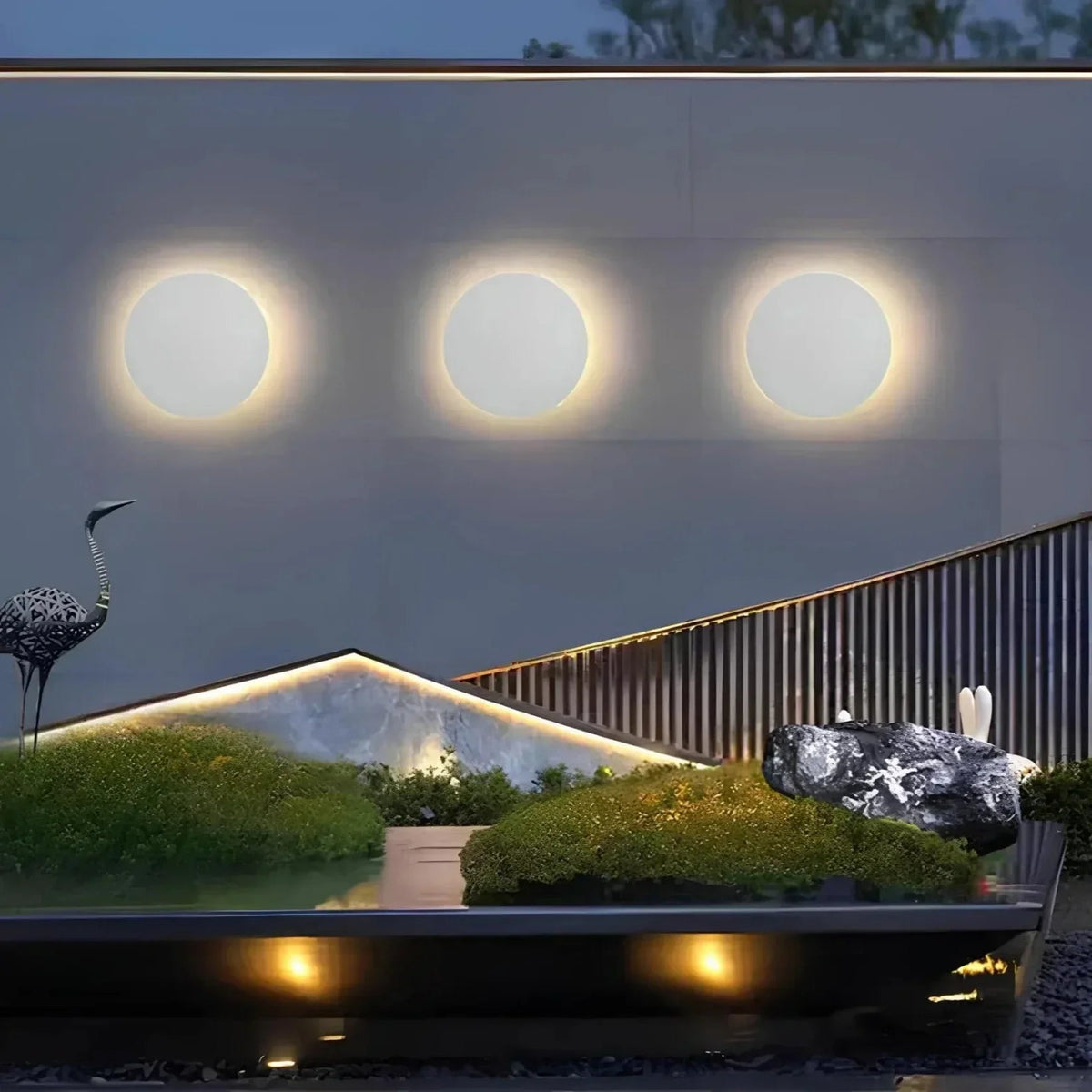 Row of three white round lights glowing on a grey outdoor wall above a landscaped garden with moss and rocks. 5