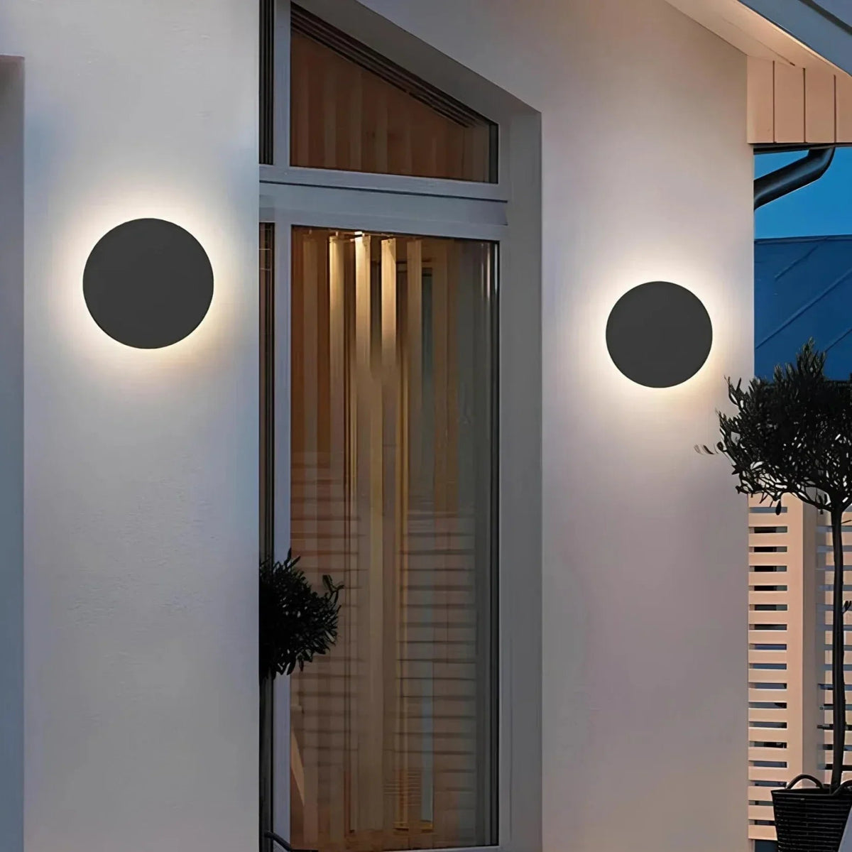 Two black round lights lit at dusk on the white exterior walls either side of a glazed front doorway. 3