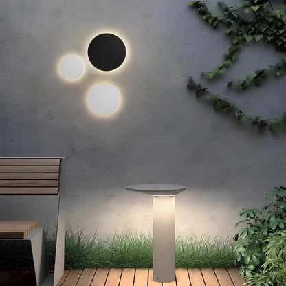 Black and white round disc lights glowing on a textured grey garden wall above a wooden deck, bench and lit bollard. 1