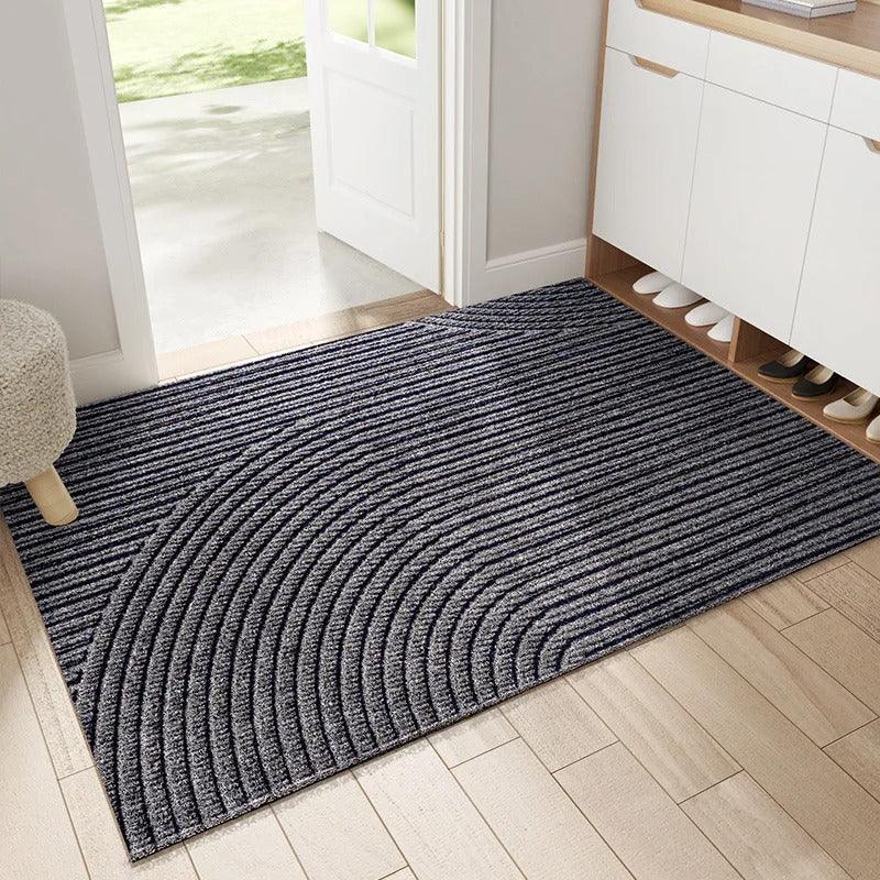 Grey and charcoal doormat with fine ridges forming sweeping curved arcs, shown in a hallway with a shoe cabinet and a stool. 2