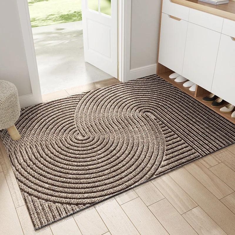Brown and beige doormat with overlapping concentric circles in raised ridges, placed on pale wooden flooring by an open doorway. 3