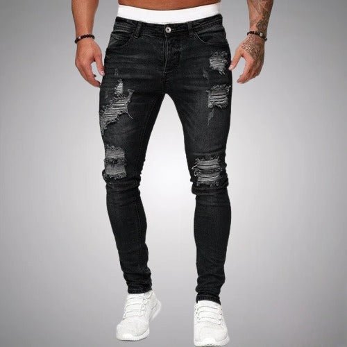 Front view of slim-fit ripped jeans in black, worn by a model with white trainers, showing frayed rips on thighs and knees. 1