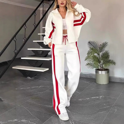 Model standing by a staircase in the white lounge set with red stripes, zip jacket worn open over a white crop top. 1