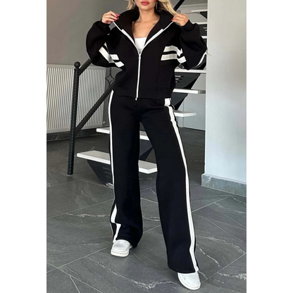Model wearing the black set with white stripes, jacket zipped up with hands at the collar, full-length view of the trousers. 3