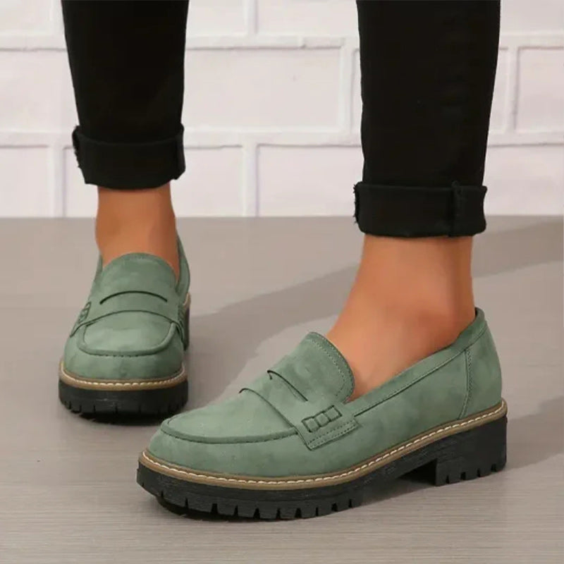 Green suede-effect penny loafers with a chunky black treaded sole, worn with rolled black trousers against a white brick wall. 1