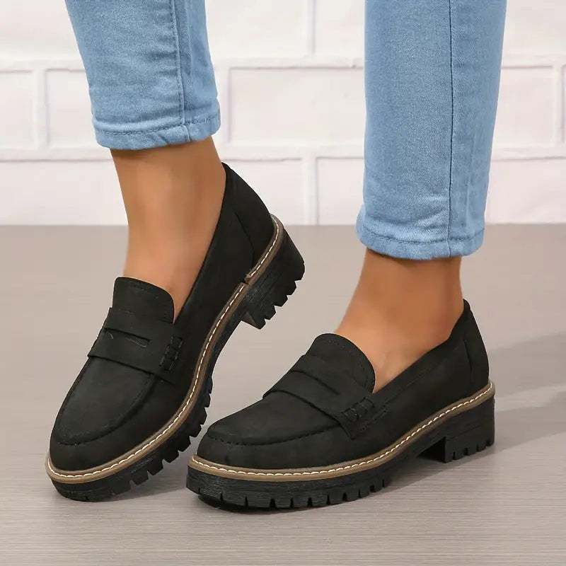 Black loafers with penny strap and thick treaded sole, worn with cuffed light blue jeans against a white brick wall. 6