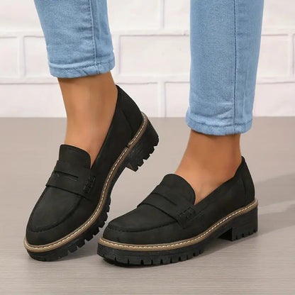 Black loafers with penny strap and thick treaded sole, worn with cuffed light blue jeans against a white brick wall. 6