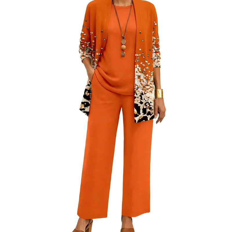 Model wearing the orange set, the long cardigan scattered with studs and a white and black speckled print towards the hem, over plain top and trousers. 3