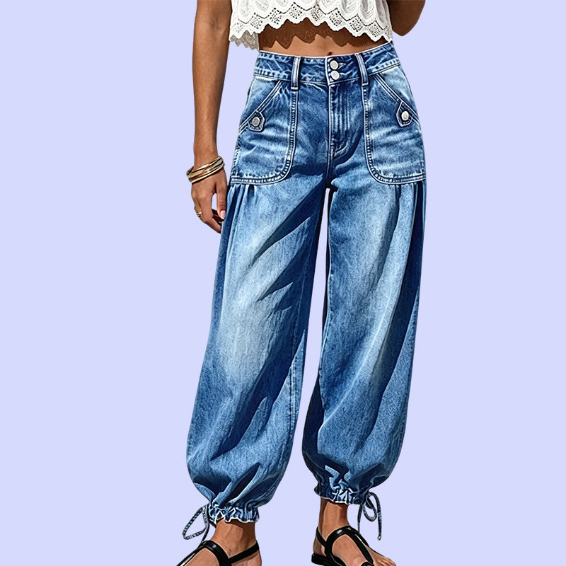 Close front view of the blue washed jeans against a lilac background, showing button-flap pockets, double-button waistband and drawstring ankle ties. 10