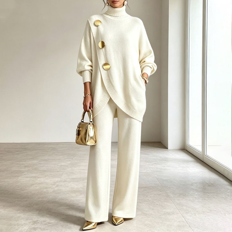Cream version of the knit set worn by a model holding a gold handbag, hand in trouser pocket. 2