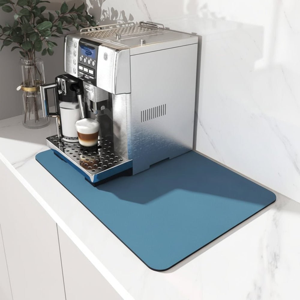 Plain blue mat under a silver bean-to-cup coffee machine on a white marble worktop, beside a glass vase with leaves. 1