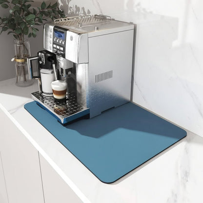 Plain blue mat under a silver bean-to-cup coffee machine on a white marble worktop, beside a glass vase with leaves. 1