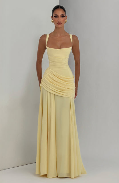 Pale yellow maxi dress worn by a model standing against a grey and white corner backdrop, front view, showing ruched hip drape. 6