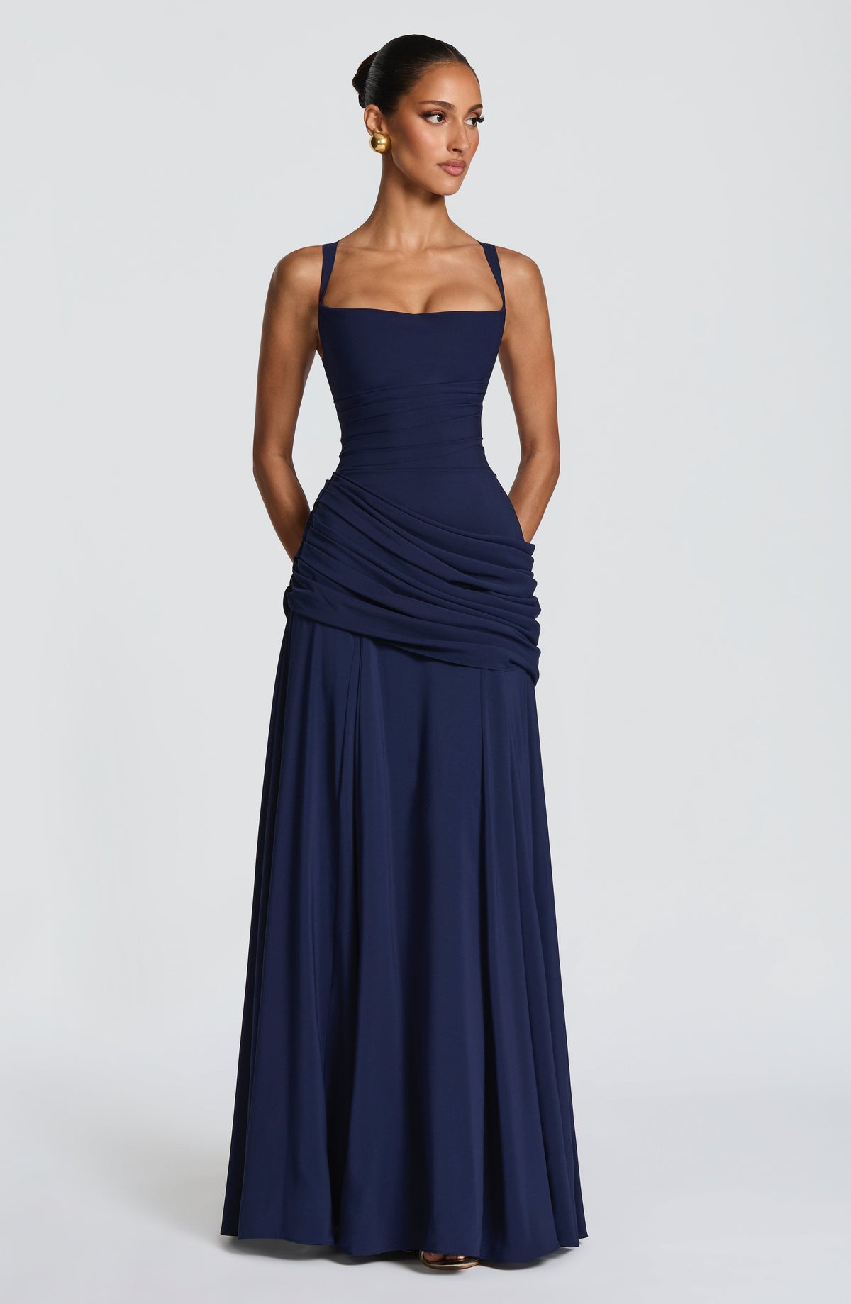 Navy maxi dress on a model looking to one side with hands in side pockets, showing the ruched hip drape and long flared skirt. 10