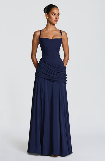 Navy maxi dress on a model looking to one side with hands in side pockets, showing the ruched hip drape and long flared skirt. 10