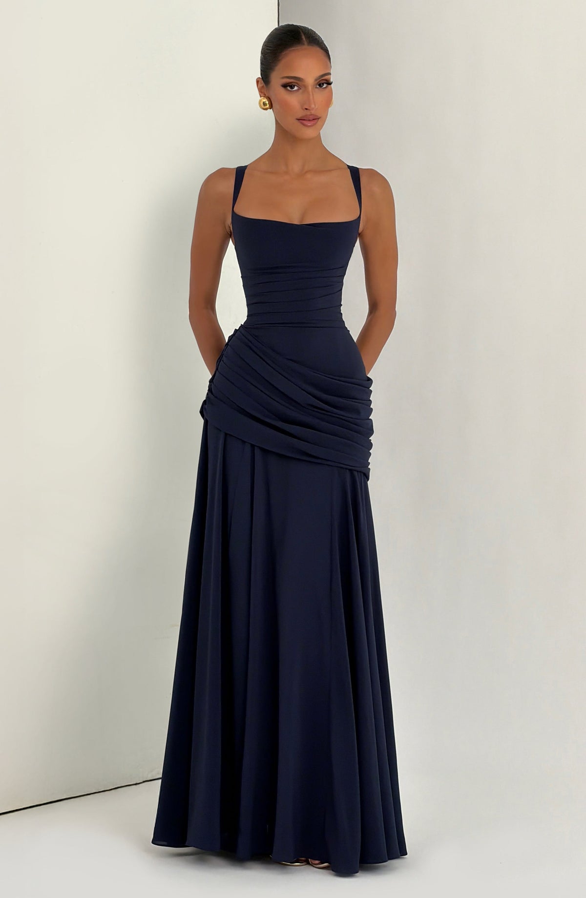 Navy maxi dress worn by a model with hands in pockets, standing against a pale grey corner backdrop, showing the draped hip and long skirt. 13