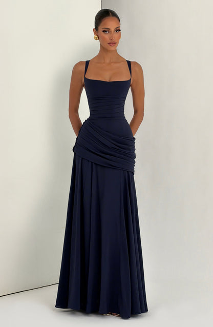 Navy dress worn by a model standing against a light two-tone wall, hands behind her, full skirt falling to the floor. 11