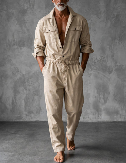 Barefoot grey-bearded model in the beige long-sleeved jumpsuit, sleeves rolled and hands in pockets, standing against a grey textured wall. 1