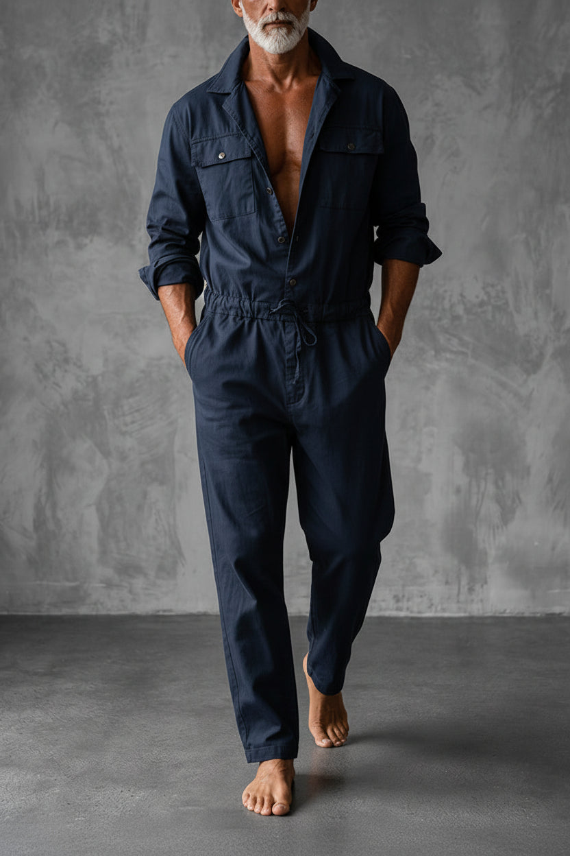Same jumpsuit in navy, worn open at the chest with rolled sleeves and a drawstring waist against a grey studio wall. 2