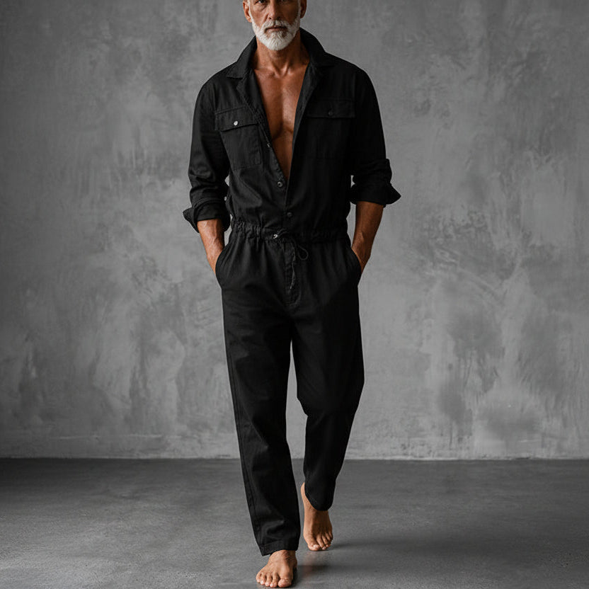 Black version of the jumpsuit on a barefoot model, with chest pockets, drawstring waist and relaxed straight legs, in a grey studio. 3