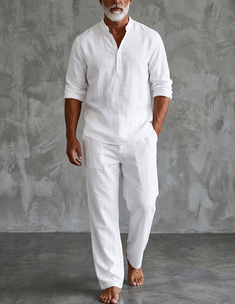 Bearded model standing barefoot against a grey textured wall in the white set, sleeves rolled and one hand in a trouser pocket. 1