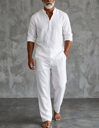 Bearded model standing barefoot against a grey textured wall in the white set, sleeves rolled and one hand in a trouser pocket. 1