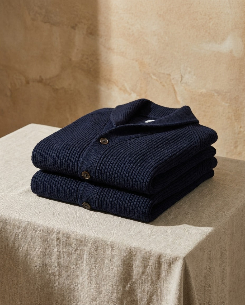 Two navy ribbed cardigans folded in a stack on a linen-covered block against a warm plaster wall, buttons and shawl collar visible. 10
